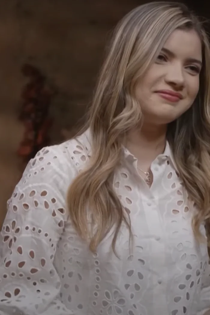 Rym Amari, cheveux blonds ondulés, chemisier blanc brodé et sourire lumineux en portrait rapproché