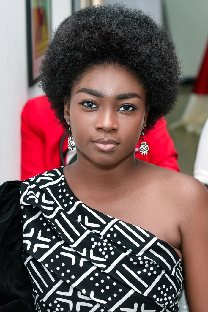 Hortavie Mpondo en portrait frontal avec afro court, boucles d'oreilles graphiques et robe noire a motifs blancs