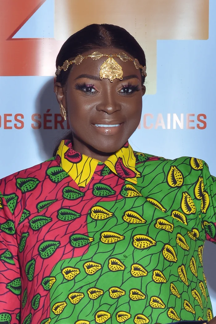 Lucie Memba souriante avec bijou frontal dore et chemise wax rouge, verte et jaune