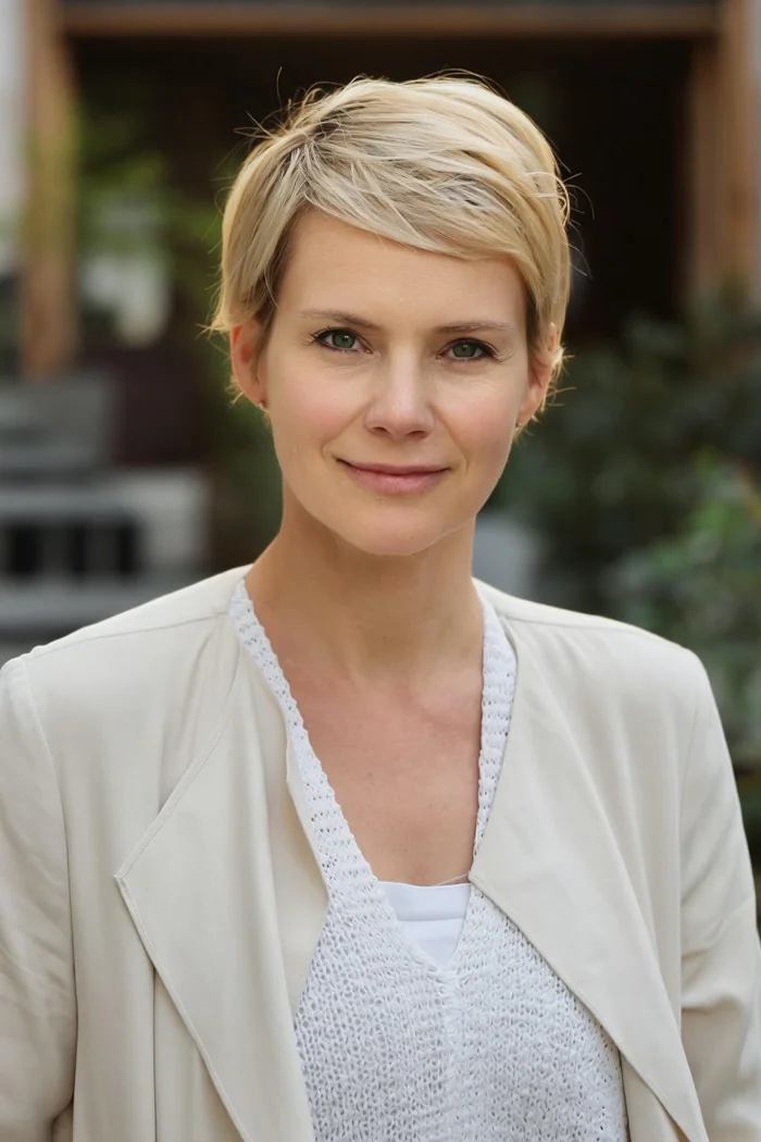 Andrea Osvárt en portrait récent, cheveux blonds courts et veste blanche devant un fond végétal flou