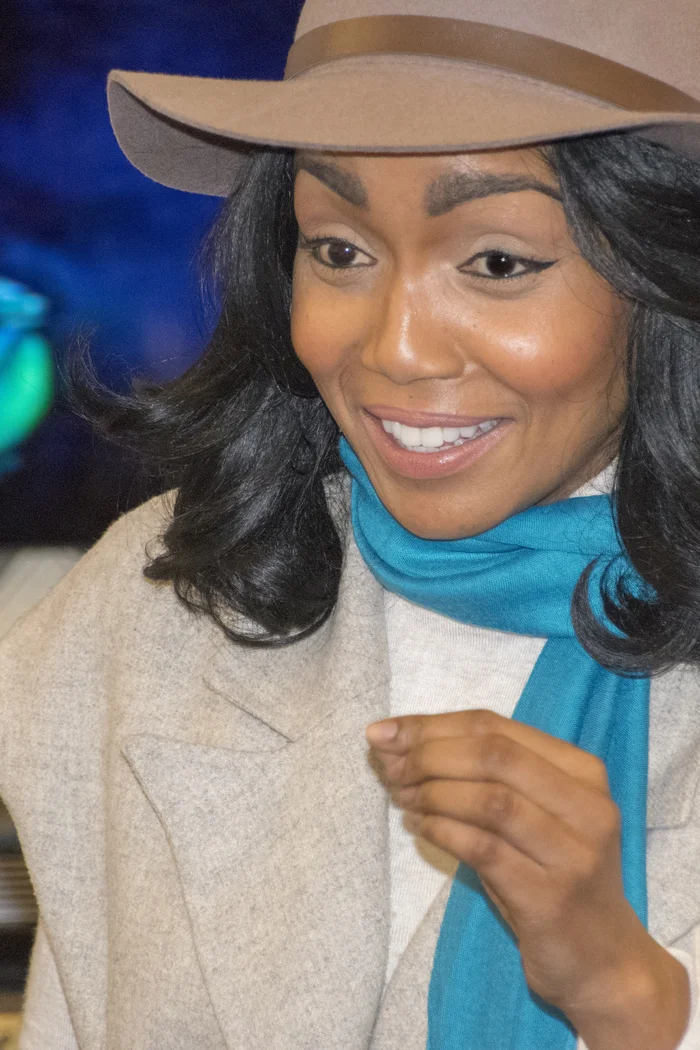 Vicky Ngari-Wilson souriante avec chapeau beige, manteau clair et écharpe bleue