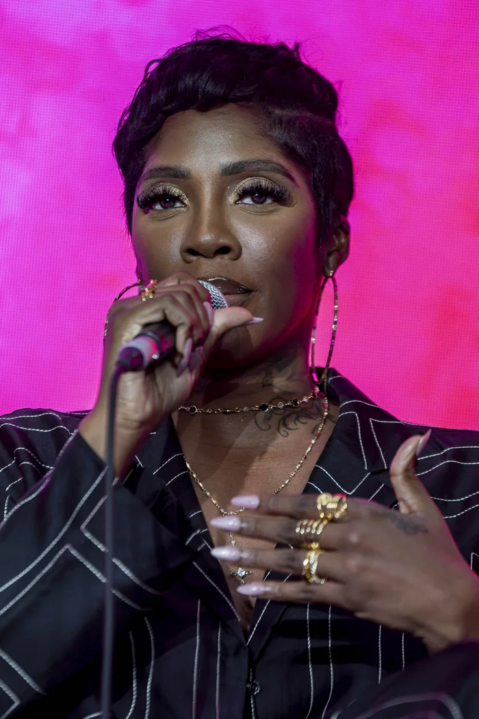 Tiwa Savage chantant devant un fond rose avec coupe courte, grandes boucles et veste noire rayee