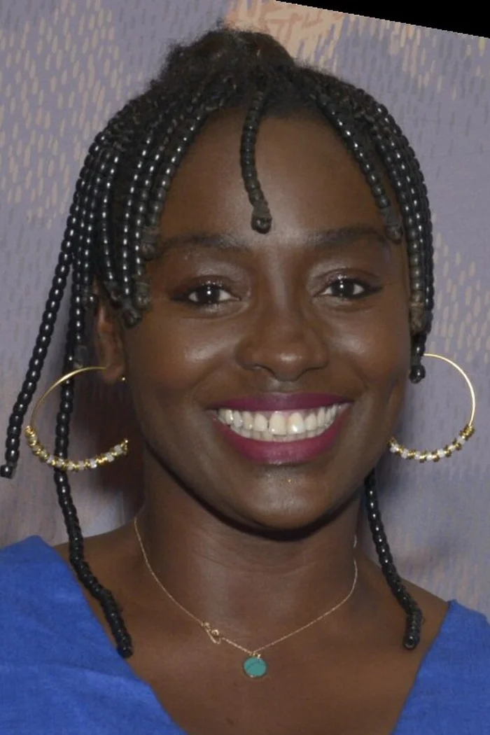 Aïssa Maïga souriante avec tresses fines, boucles d'oreilles dorées et haut bleu