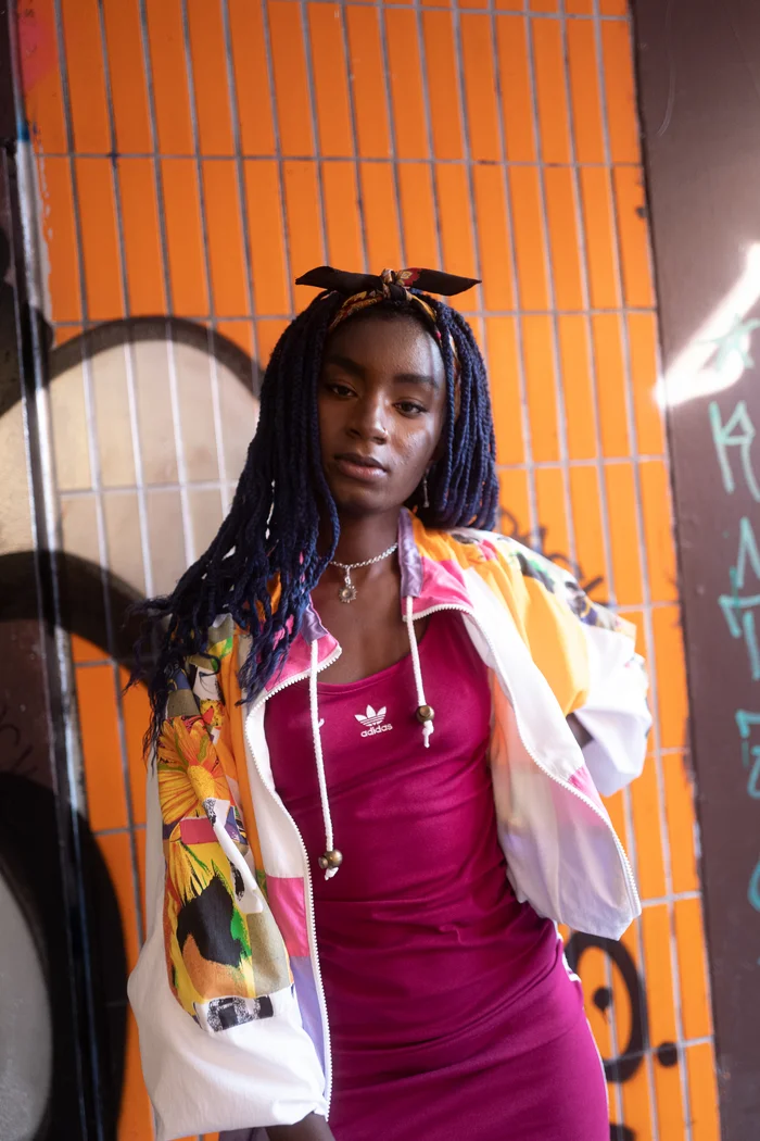 Oumi Janta en robe rose et veste wax colorée devant un fond urbain orange