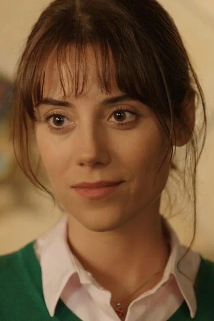 Cansu Dere souriante, cheveux bruns avec frange, chemise blanche et cardigan vert.