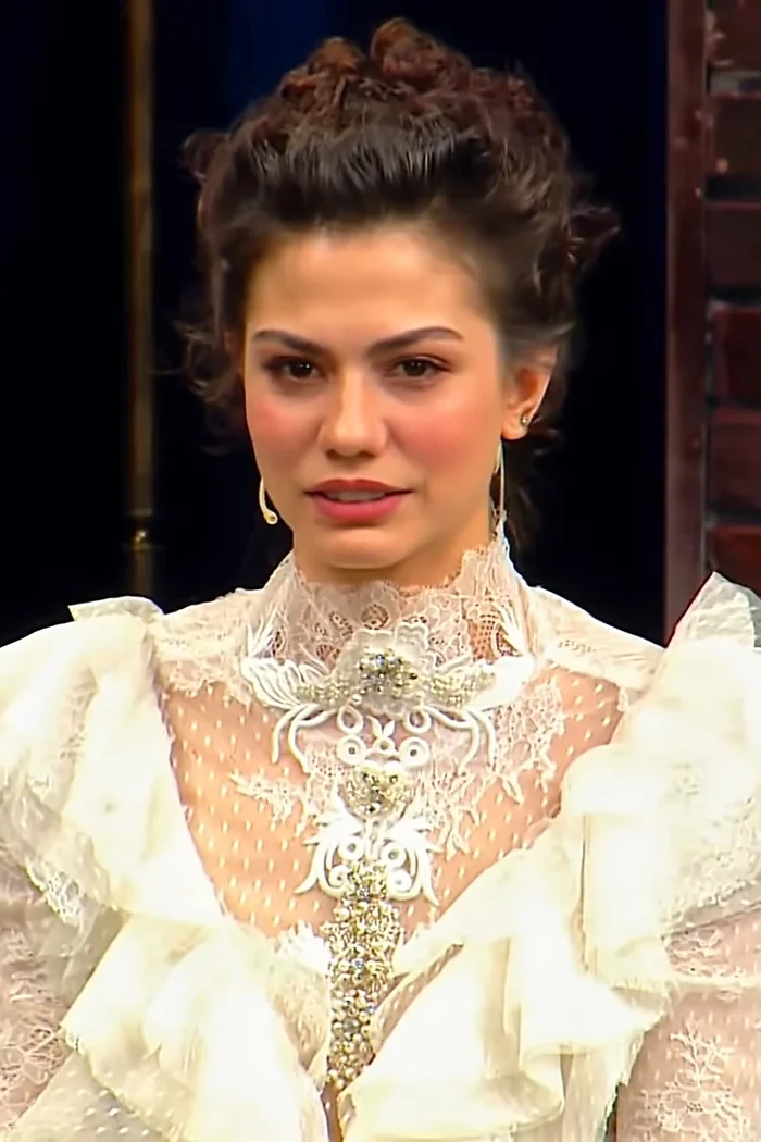 Demet Özdemir, coiffure relevée, robe blanche en dentelle et bijoux sur fond sombre.