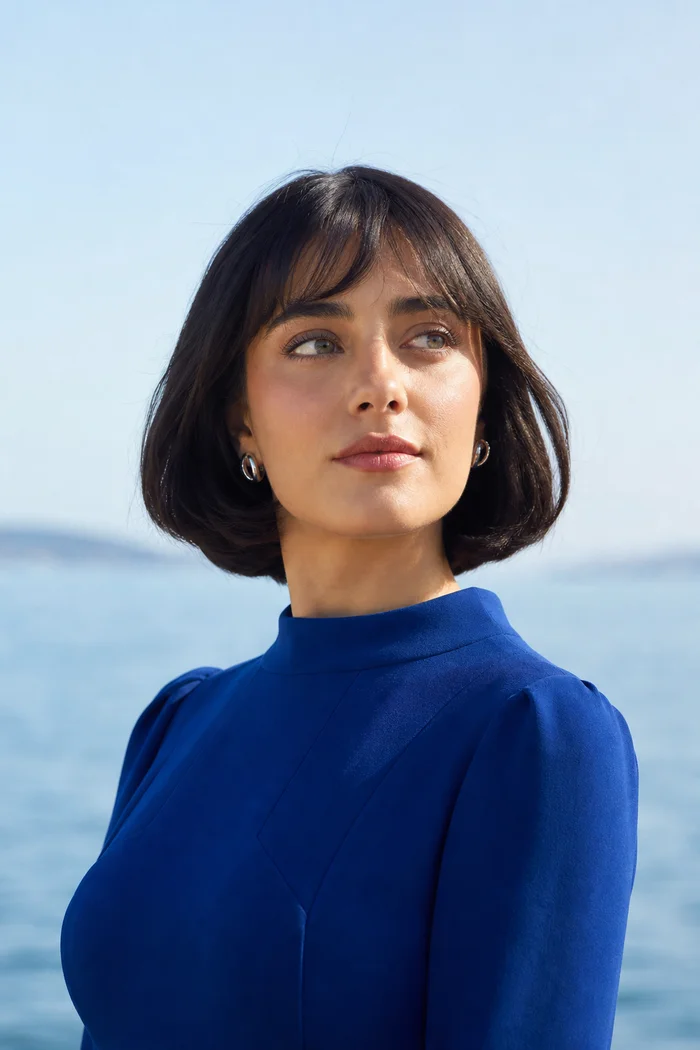 Femme fictive à la coupe brune avec frange, robe bleu cobalt et yeux gris-bleu devant une mer lumineuse.